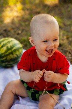 Joyful baby in watermelon outfit laughing outdoors with a whole watermelon nearby