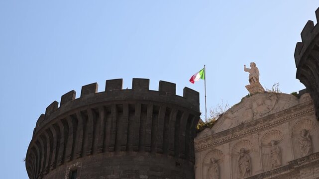 Italian flag ripples in the breeze above the stone tower and statues of Castel Nuovo against a clear blue sky.
