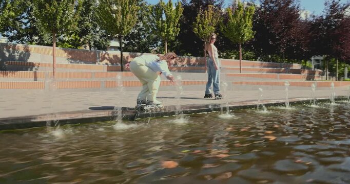 Two young women rollerblade beside urban fountains as side tracking shot captures friendship and playful movement. Summer recreation, active lifestyle and social connection concept.