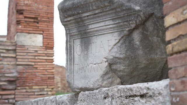 Cracked Latin inscription block rests on a ledge between faded brick structures in a static daytime shot.
