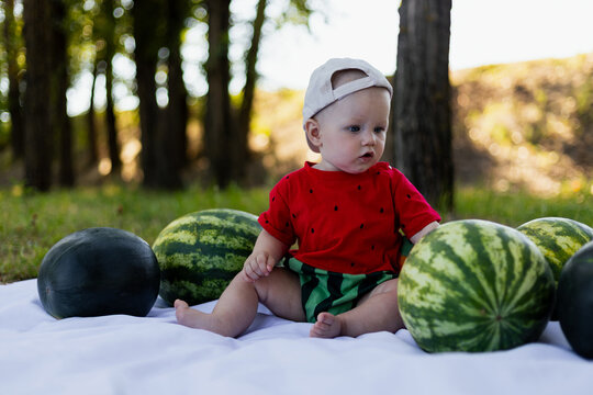 Adorable baby in watermelon outfit sits with watermelons in a park