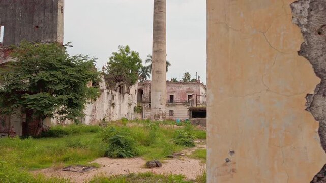 Historic ruins featuring weathered stone walls and a tall column surrounded by an overgrown courtyard. Palm trees, grass, and deteriorating old buildings create a weathered archaeological setting