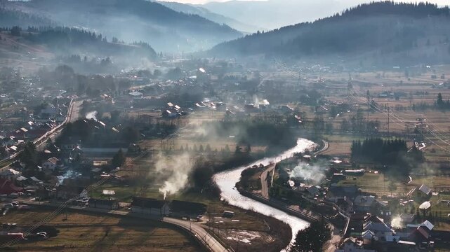 Elevated winter view of the Dorna River winding through houses and rural neighborhoods near Vatra Dornei, Romania, with smoke rising from chimneys and mountains in the background.
