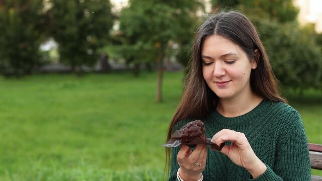 Young woman opens chocolate cupcake wrapper and eats cupcake. She calmly enjoys sweet food in park wearing green sweater.