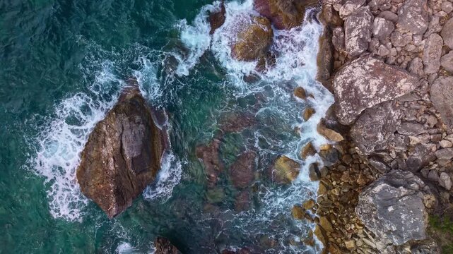 Aquarium Bay Albania shoreline fills view as waves break over rocks. Aerial perspective reveals teal water, foamy surf, and scattered boulders along rugged coast, creating calm yet powerful mood.