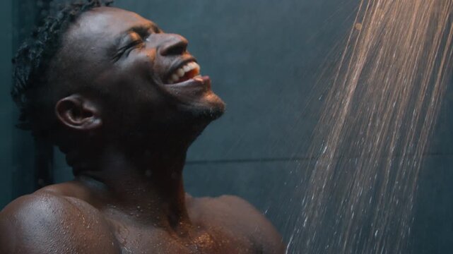 Happy African American muscular man dancing and singing in shower under hot falling water naked wet ethnic guy athletic male listening music washing body funny dance in dark bathroom morning routine