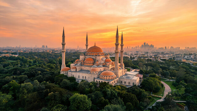 A grand historic mosque with multiple domes and minarets stands amidst greenery, with a city skyline at sunset in the background.