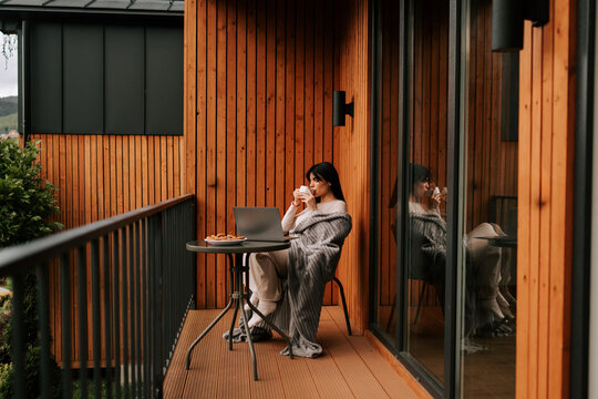 Pregnant woman working on a laptop with a cup of coffee on the balcony of a cozy wooden cottage