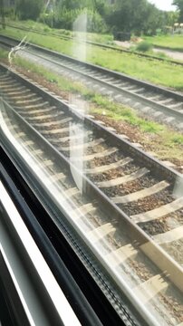 Parallel steel rails and concrete sleepers stretch across gravel ballast beside lush summer greenery through a passenger window. Gentle reflections glide over the pane in bright daylight.