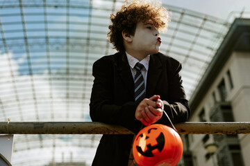 Fototapeta premium Caucasian child wearing Halloween costume makeup leaning on railing outdoors holding pumpkin bucket. Image suitable for seasonal holiday marketing and trick or treat event promotion