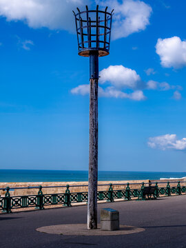 Historical maritime fire beacon basket on tall wooden pole at seaside promenade under blue sky