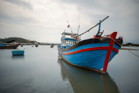 Traditional blue and red wooden fishing boat rests moored in calm Vietnamese harbor under cloudy sky. Round basket boat and smaller vessels float near coastal village with houses and low green hills.