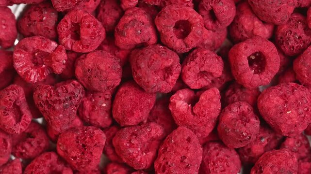 Rotating freeze-dried raspberry pieces shot in top-down macro close-up under bright studio lighting. Crisp red textured berry pieces fill the entire frame