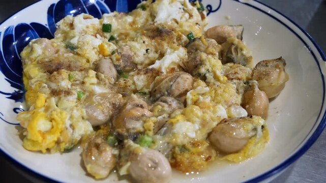 Thai oyster omelet with fresh oysters scrambled in fluffy egg and spring onion on blue and white porcelain plate, delicious seafood street food dish close-up