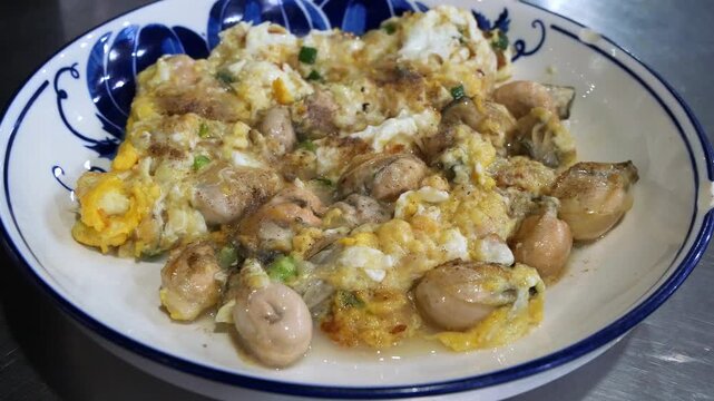 Thai oyster omelet with fresh oysters scrambled in fluffy egg and spring onion on blue and white porcelain plate, delicious seafood street food dish close-up