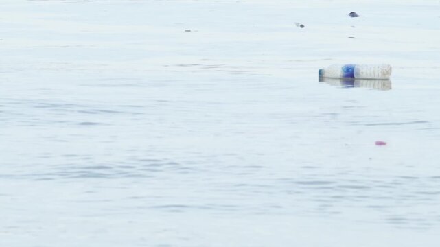 Discarded crumpled plastic bottle with green cap floating on calm pale blue water, river pollution and marine plastic waste environmental problem concept with copy space