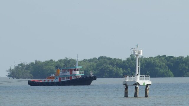 Colorful tugboat towing a small fishing boat with rope across calm blue sea, green navigation buoy and seagull on horizon