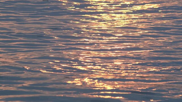 Golden sunset light reflecting and sparkling on gentle blue water ripples, warm shimmering path across calm river surface, tranquil evening footage
