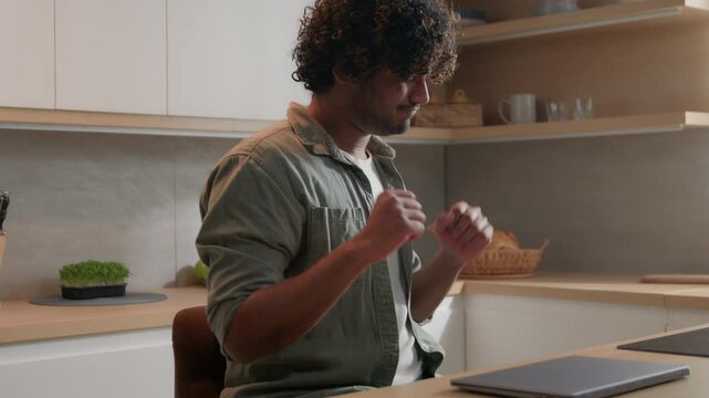 Active Indian Hispanic Arabian man dancing at kitchen table with closed laptop happy freelancer remote worker celebrating day off finish work joyful student businessman male funny movement at home