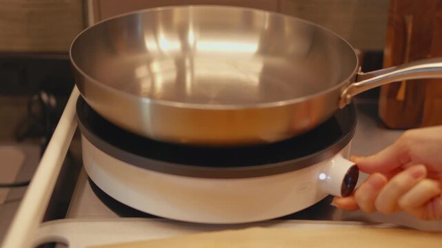 Hand placing stainless steel frying pan on portable induction cooktop and adjusting temperature knob. Close-up of heating up skillet on tabletop stove in home kitchen for cooking prep process.