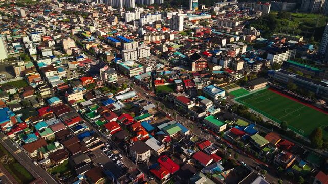 Aerial forward flight over dense residential neighborhood of Geumsan town in Chungcheongnam-do, South Korea, with colorful rooftops, narrow streets, school buildings and green soccer field