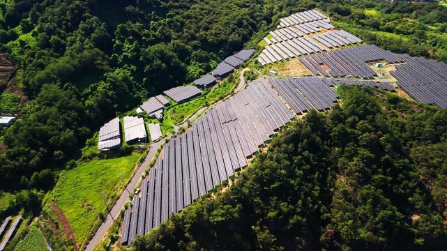 Aerial parallax across large solar farm cut into forested hillside in Geumsan South Korea where dark photovoltaic arrays and canopy roofed structures follow curving access road down slope