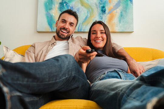 A cheerful couple relaxes on a sunny yellow sofa, sharing a remote while looking toward a television. The close framing highlights connection, comfort, and everyday home entertainment in a warm moment