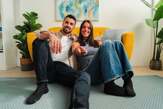 A joyful couple relaxes on a yellow sofa, smiling at the camera, sharing a light moment of connection and closeness. This intimate portrait captures warmth, friendship, and everyday comfort in a
