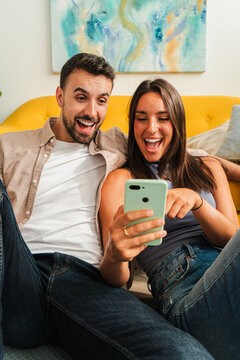 A joyful couple relaxes on a sofa, sharing a moment of amusement as they view content on a smartphone, conveying warmth, connection, and lighthearted fun in a living room setting.