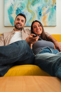Two smiling adults sit close on a bright yellow sofa, holding a remote control and enjoying a shared moment of entertainment. The scene conveys warmth, connection, and relaxed home comfort.