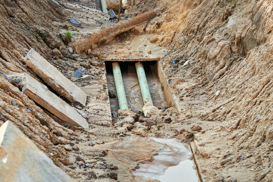 Deep excavated trench reveals two green pipes running through concrete casing at construction site during municipal utility repair project. Excavated trench, exposed pipes laid in concrete channel