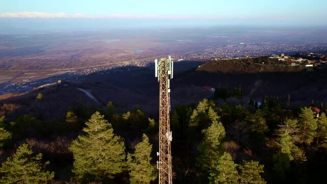 Tall telecom mast dominates the foreground above pine trees and sections of a stone defensive wall while patchwork fields a valley village and the snowy Caucasus span the wide Georgian panorama
