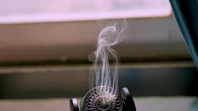 Close-up of a black electronic aroma diffuser emitting a swirling plume of white mist. The modern device sits indoors near a bright window and dark curtain during daytime, creating a relaxing mood.