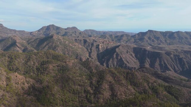 A majestic aerial drone push in shot reveals the vast, rugged, and volcanic mountainous landscape on Gran Canaria island, Spain. Pine forests dot the slopes leading down to the distant Ocean.