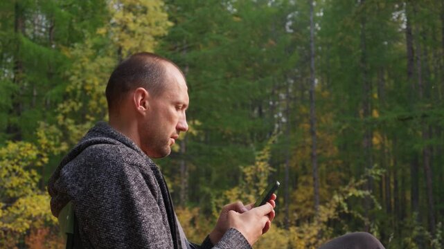 A man in a gray hoodie sits in an autumn forest, intently using his smartphone with minimal movement, surrounded by trees with green and yellow leaves under soft, diffused daylight.