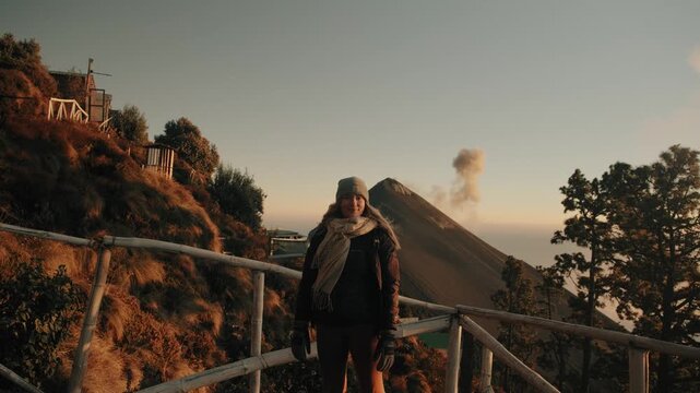 Young female hiker stands bundled at dawn on Acatenango campsite, gazing at active Fuego volcano erupting amid golden sunrise, rustic fencing, scenic mountains, and dramatic warm light.
