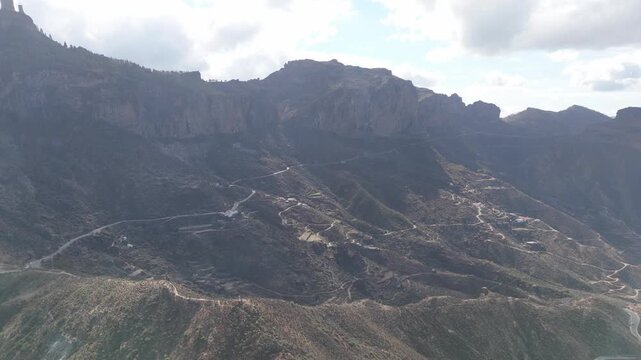 Aerial pan right drone shot over the stunning, rugged Tejeda mountains on Gran Canaria, showcasing winding roads, small mountain villages, and dramatic cliff faces under an overcast sky.