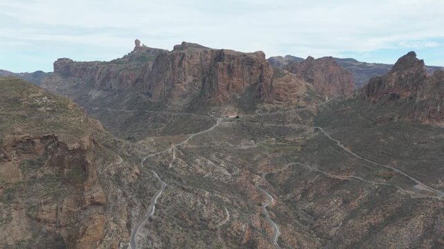 Aerial drone footage pans right over the rugged, volcanic landscape of Gran Canaria, Canary Islands. Winding roads cut through deep valleys and reveal the iconic Roque Nublo.