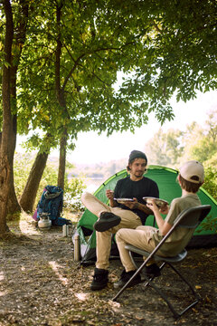 Father and son eat from metal plates on camp chairs beside tent. Family meal, outdoor living and camping comfort concept.
