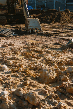 Excavator bucket resting on muddy uneven ground at an active construction site, surrounded by excavated soil, puddles and construction materials during earthwork and site preparation.