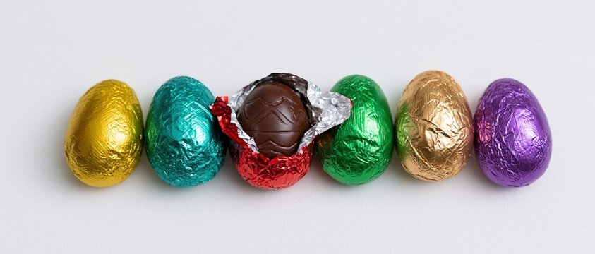 Six foil-wrapped chocolate easter eggs in a row on white background, one unwrapped revealing chocolate interior