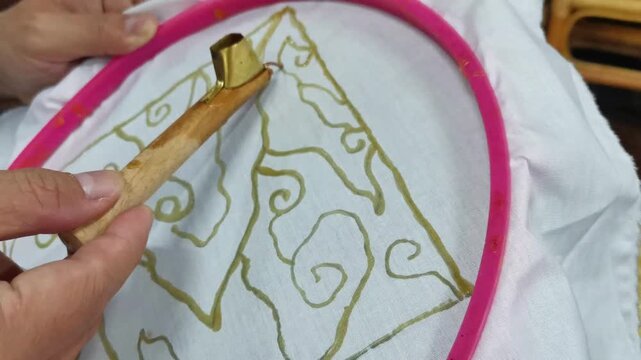 Hands holding traditional canting tool to apply hot wax on fabric for indonesian batik making