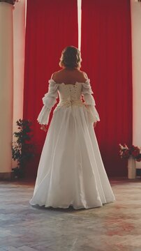 Sexy woman queen opens red curtains throws it to the sides, uncovers window looking out enjoy day sun light. Happy girl princess blonde hair white vintage dress old style back rear view vertical video