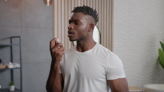 Sad African American man with bad breath smell spraying refreshing spray freshener in mouth at home bathroom smiling male guy applying oral hygiene dental care product fresh breathing morning routine
