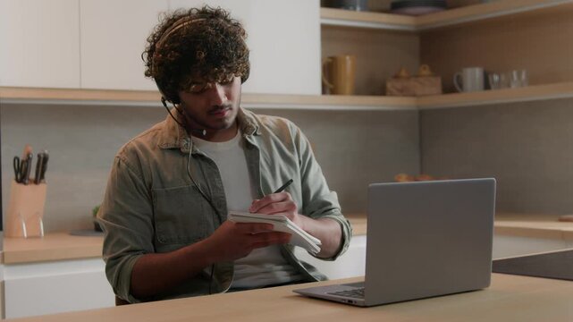 Indian Arabian student man wearing earphones studying online at home kitchen watching laptop lecture video call ethnic male in headset writing notes remote education course distance learning indoors