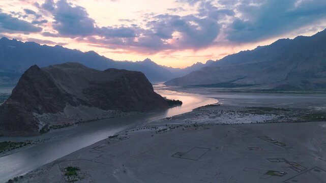 Aerial forward and upward flight over winding Indus river in Skardu valley desert landscape at sunset, wide river snaking through arid mountain valley under sunset sky