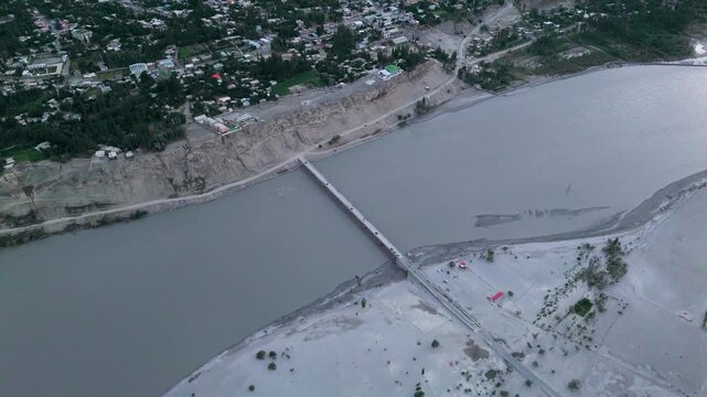Aerial forward tracking shot over bridge crossing grey waters of Indus River connecting town and sandy riverbank, traffic crossing long bridge over wide river towards mountain valley settlement