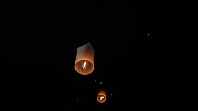 Warm glowing paper sky lantern rising at night with visible flame inside glowing thin paper and blurred lanterns in outdoor evening sky