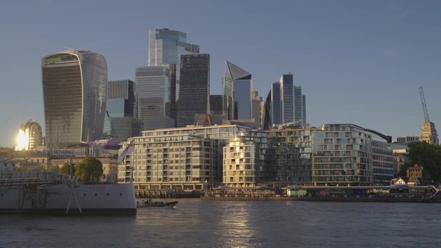 Medium low-angle view across the River Thames highlighting the modern skyscrapers of the City of London and HMS Belfast in warm evening sunlight.