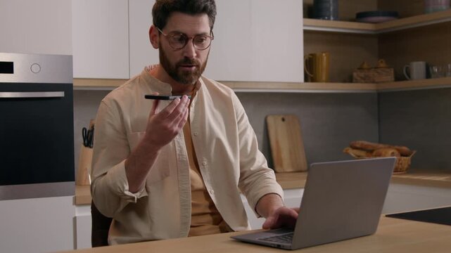 Multitasking Caucasian business man at kitchen working laptop recording voice message mobile phone remote focused busy freelancer in glasses businessman guy male speaking telephone AI app indoors